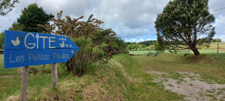 Panneau peint en bleu indiquant l'entrée du gîte Les Petites Poules de Kerbiguet sur un chemin en terre